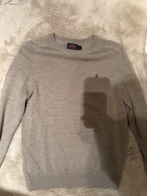 Ljusgrå merinoulltröja Morris - Säljer en stilren ljusgrå tröja från Morris i mjuk merinoull. Tröjan har rund halsringning, diskret broderad logga på bröstet och långa ärmar. Perfekt för lager på lager under kyliga dagar. S passar M
