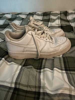Nike Air Force 1 vita sneakers - Säljer ett par klassiska Nike Air Force 1 sneakers i helvitt med snörning och perforerad tå. Skorna har en platt sula och rund tå, tillverkade i skinn med textilfoder. Perfekt för dig som gillar tidlös streetstil och vill ha ett par ikoniska sneakers.