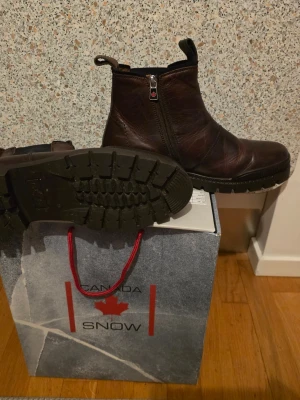 Canada Snow Chelsea boots brun stl 42 - Säljer ett par snygga Chelsea boots från Canada Snow i mörkbrunt skinn. Skorna har grov platt sula, dragkedja på ena sidan och elastisk panel på andra sidan för enkel påtagning. Perfekt för dig som gillar robust stil och vill ha något som funkar hela vintern.
