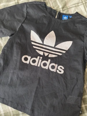 Svart mesh t-shirt från Adidas - Säljer en svart t-shirt från Adidas med det klassiska trefoil-loggan i vitt framtill. Tröjan är gjord i meshmaterial med små hål över hela och har korta ärmar. Baksidan har en diskret dragkedja upptill för enkel på- och avtagning. Perfekt för dig som gillar sportig och streetwear-stil.