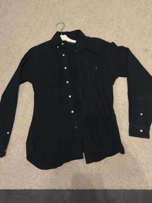 Mörkblå skjorta Ralph Lauren Slim Fit - Klassisk mörkblå skjorta från Ralph Lauren i slim fit-modell. Skjortan har vita knappar, lång ärm och den ikoniska röda loggan broderad på bröstet. Tillverkad i mjuk bomull och passar perfekt till både jeans och chinos. Enda deffekten är att knappen löngst ner har gått av men går lätt att sy på en ny, kan skicka med extra knapp! Passar för både S/M