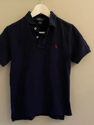 Mörkblå polo t-shirt från Ralph Lauren - Snygg mörkblå polo t-shirt från Ralph Lauren med klassisk krage och två knappar framtill. Säljer då den är för liten. Den är i storlek 10-12 år. Hör av er vid funderingar 