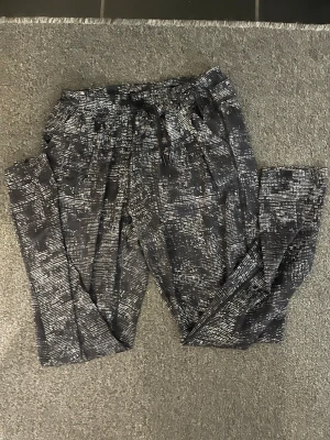 Svarta mönstrade träningsbyxor med snörning - Säljer ett par svarta träningskläder med vitt abstrakt mönster och snörning i midjan. Byxorna har en loose passform och är perfekta för chill dagar. Materialet känns mjukt och skönt mot huden. Snygga att matcha med en enkel t-shirt eller hoodie.