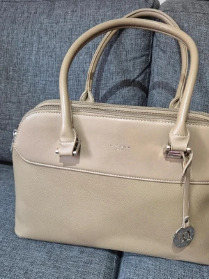 Handväska från David Jones - Stilren beige eller khaki handväska från David Jones i skinnimitation med två rejäla handtag och silverfärgade metalldetaljer. Väskan har flera fack med dragkedja och en rund metallbricka med logga som detalj. Perfekt för dig som vill ha ordning på dina grejer och gillar klassisk design.🛍🫶🏻