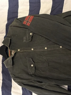 Grön manchester skjorta Napapijri - Snygg grön skjorta från Napapijri i mjuk manchester med två bröstfickor och vita knappar. Långärmad modell med coolt norskt flaggmärke på ena ärmen och detaljer vid kragen. 