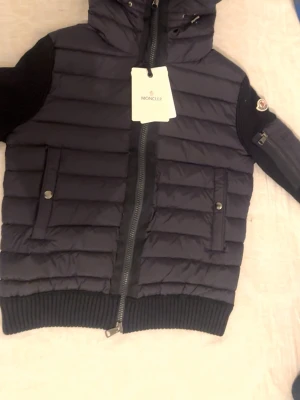 Svart pufferjacka från Moncler - Snygg svart pufferjacka från Moncler med huva och ribbade muddar. Jackan har quiltad design, dragkedja framtill och två fickor med tryckknappar. Ärmarna är stickade och har en Moncler-logga på ena armen. Perfekt för kalla dagar.