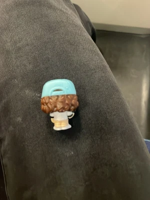 Vit väst med blå detaljer - Säljer en liten vit väst med blå detaljer som syns på en figur med brunt lockigt hår och en blå keps med texten 'Thinking Cap'. Västen har en enkel design och är gjord i syntetmaterial, perfekt för dig som gillar unika accessoarer till samlarfigurer. 