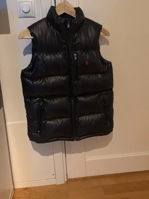 Svart dunväst från Polo Ralph Lauren - Säljer en svart dunväst från Polo Ralph Lauren med hög krage och dragkedja framtill. Västen har två sidofickor med dragkedja och en bröstficka, samt den klassiska röda loggan broderad på bröstet. Perfekt för lager på lager under höst och vinter.