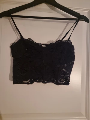 Svart spetsig croppad linnetopp - Säljer en svart croppad linnetopp från Gina Tricot med tunna axelband och vacker spets över hela toppen. Toppen har en feminin och elegant look med genombruten spets och lätt vågig kant nedtill. Perfekt att styla till jeans eller kjol.