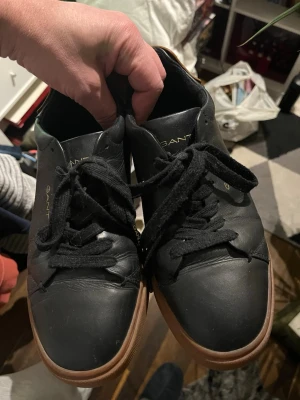 Svarta sneakers från GANT, strl 41 - Snygga svarta sneakers från GANT med bruna gummisulor och svart snörning. Skorna är i skinn med rund tå och har GANT-logga i guld på sidan och plösen. Perfekta för dig som gillar stilrena och klassiska sneakers.