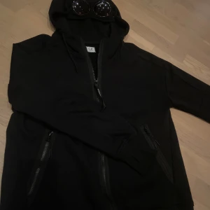 Svart C.P. Company hoodie med goggles - Svart hoodie från C.P. Company med ikoniska goggles i huvan. Tröjan har dragkedja framtill, två fickor med dragkedja och långa ärmar. Tillverkad i mjuk bomull och har en clean, urban look som sticker ut. Tröjan är använd 1-2 gånger, då den är för liten och har inte kvar kvittot så säljer den där med här.