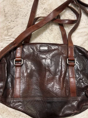 Brun axelväska från Spikes & Sparrow - Snygg brun axelväska i skinn från Spikes & Sparrow med justerbar rem och coola metalldetaljer. Väskan har ett stort huvudfack med dragkedja, flera innerfickor och en praktisk ficka med spänne på framsidan. Perfekt för dig som gillar klassisk stil med lite edge.