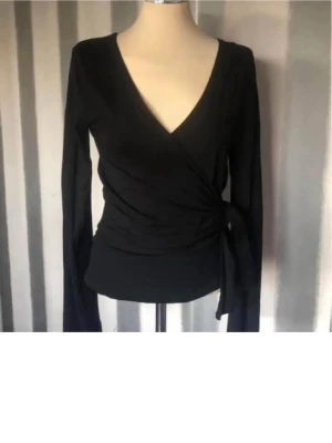 Svart omlottopp med knyt - Säljer en svart omlottopp med lång ärm och v-ringad hals. Toppen har knyt i sidan som ger en snygg siluett och är tillverkad i ett mjukt, stretchigt material. Perfekt att styla med jeans eller kjol för en cool look.