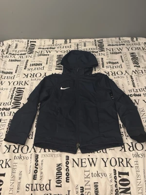 Mörkblå Nike vindjacka med huva - Säljer en mörkblå Nike Shield vindjacka i storlek M 137- 147. Jackan har huva, dragkedja framtill och två sidofickor med dragkedja. Tillverkad i lätt och tålig polyester, perfekt för blåsiga dagar. Klassisk Nike-logga på bröstet och justerbara ärmslut.