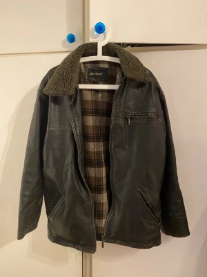 Svart skinnjacka med teddykrage Janbell - Säljer en svart skinnjacka från Janbell med mjuk teddykrage och rutigt innerfoder. Jackan har dragkedja framtill, tre fickor och justerbara spännen bak. Perfekt för dig som vill ha en klassisk look med lite edge.