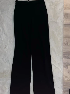 Svarta kostymbyxor från Vero Moda - Snygga svarta kostymbyxor från Vero Moda med raka ben och hög midja. Byxorna har bälteshällor, diskret dragkedja och knapp framtill. Perfekta till en stilren look och passar till allt. Materialet är mjukt och följsamt, vilket gör dem riktigt bekväma.  (Regular i längden)