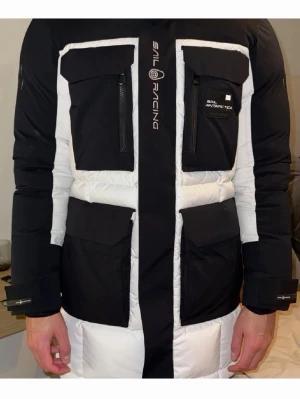 Sail Racing svartvit dunjacka - Säljer en fet svartvit dunjacka från Sail Racing med stora fickor framtill och dragkedjor. Jackan har lång ärm, hög krage och coola detaljer med logga på bröstet. Perfekt för kalla dagar när du vill hålla stilen. Jackan har 12 fickor!