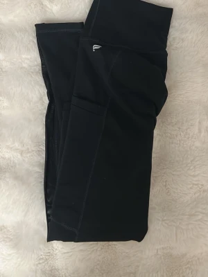 Träningstights med ficka  - Svarta träningstights från Fabletics med PowerHold-teknologi. Hög midja och ficka på sidan till mobil, perfekta för gymmet eller löprundan. Snygg och enkel design med diskret logga på höften. Säljer då jag har alldeles för många träningstights. Bara testade. 