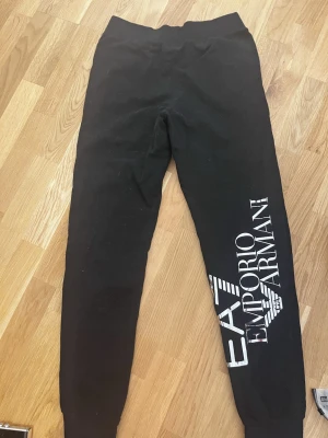 Svarta mjukisbyxor från Emporio Armani - Svarta mjukisbyxor från Emporio Armani med stort vitt tryck längs ena benet. Byxorna har resår i midjan och muddar vid bensluten för en skön och avslappnad passform. Perfekta för chill eller streetwear. Oanvända pga passform 