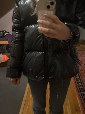 Svart glansig pufferjacka - Säljer en svart, glansig pufferjacka med dragkedja framtill. Jackan har en oversized passform och långa ärmar. Materialet är syntet och ger en riktigt modern vibe. Perfekt för kalla dagar när du vill vara både varm och snygg.