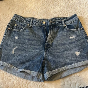 Blå denimshorts med slitningar - Snygga blå jeansshorts med rullade benslut och coola slitningar framtill och baktill. Klassisk femficksmodell med knapp och dragkedja. Perfekta för sommarens alla äventyr, loose fit och skön passform.