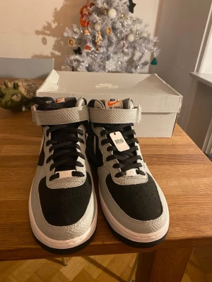 Nike Air Force 1 Mid '07 svart/grå - Säljer ett par Nike Air Force 1 Mid '07 i svart och grått med cool textur på ovandelen. Skorna har klassisk snörning, kardborreband vid ankeln och vit mellansula. Perfekt för dig som vill ha en ikonisk sneaker med snygga detaljer.