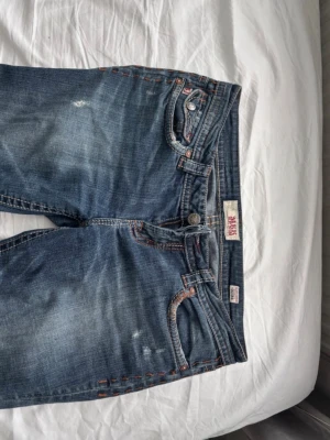 Blå bootcut jeans  - Säljer ett par blå jeans med bootcut passform och låg midja. Jeansen har snygga slitningar, kontrastsömmar och broderade bakfickor med silverfärgade knappar. Materialet är klassisk jeans i bomull med detaljer som ger extra edge.midja 43cm och innerben 84cm. de är lite slitna längst ner bak på benen men inget man egentligen stör sig på💕
