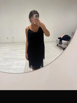 Svart fransklänning med smala axelband - Säljer en svart klänning med smala axelband och v-ringning. Klänningen har fransdetaljer som ger en snygg och rörlig look. Modellen är figurnära och slutar strax ovanför knäna. Perfekt för dig som vill sticka ut med en klassisk men trendig stil.