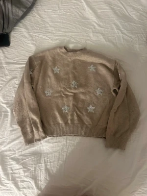 Beige stickad tröja med stjärnor Gina Tricot - Beige stickad tröja från Gina Tricot med silverfärgade paljettstjärnor framtill. Tröjan har ribbade muddar vid ärmslut och nederkant samt rund hals. Perfekt för dig som vill ha en mysig och snygg vardagslook.