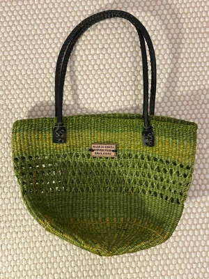 Grön shoppingväska i sisal - Säljer en handgjord shoppingväska som ja köpte i Kenya i flätad sisalfiber. Väskan är grön med inslag av gult och har svarta handtag i syntet. Perfekt storlek för att bära med sig allt man behöver och har en luftig, öppen väv som ger en chill vibe. Aldrig använt