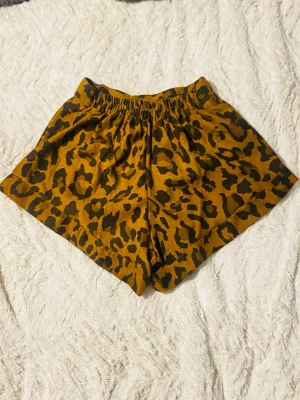 Leopardmönstrade shorts med knapp - Säljer ett par bruna shorts med leopardmönster och svarta fläckar. De har elastisk midja bak och en knapp framtill för extra detalj. Materialet känns mjukt och luftigt, perfekt för varma dagar. Passformen är loose och de har en avslappnad vibe.  Storlek: M