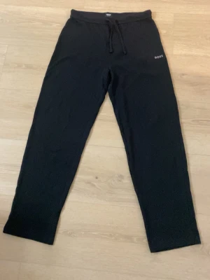 Svarta mjukisbyxor från BOSS, stl - Svarta mjukisbyxor från BOSS med dragsko i midjan och diskret logga på vänster höft. Byxorna har en rak passform och är tillverkade i mjukt material som känns skönt mot huden. Perfekta för chill eller träning.