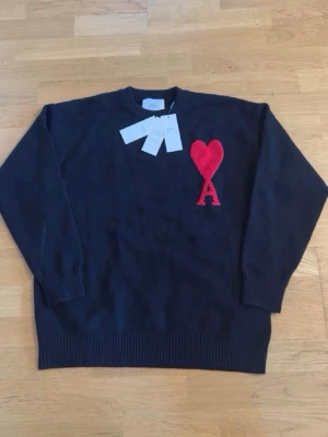 Mörkblå stickad tröja från Ami Paris - Sprillans ny stickad tröja från Ami Paris med rund hals och ribbade muddar. På bröstet finns ett stort rött hjärta med bokstaven A. Perfekt för dig som gillar stilrena plagg med en twist. Materialet är mjukt och skönt.