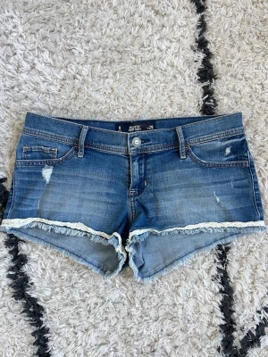 Blå low rise shorts Hollister - Snygga blå jeansshorts från Hollister i storlek w29, short short med låg midja, slitna detaljer och råa kanter. Shortsen har vit spets längs bensluten och klassiska fickor både fram och bak. Midjemått 43cm mätt rakt över 💙