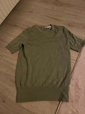 Olivgrön stickad topp från ZARA - Olivgrön stickad topp från ZARA med korta ärmar och rund halsringning. Toppen har ribbade muddar och tre dekorativa knappar i nacken där bak. Perfekt till jeans eller kjol för en chill look.köpte för 300 men säljer för 100