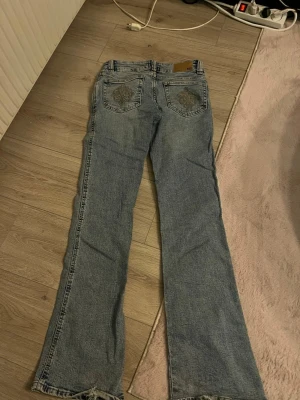 Blå bootcut jeans med broderade fickor - Säljer ett par ljusblå bootcut jeans med snygga broderade detaljer på bakfickorna. Jeansen har klassisk femficksdesign och är tillverkade i jeansmaterial. Perfekta för dig som gillar en lite retro vibe och vill ha något som sticker ut. köpte för 400kr. säljer för 300 men skriv  så kanske vi kan gå ner i pris.