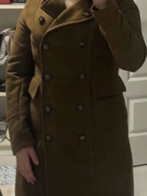 Brun kappa från H&M, strl 34 - Jackan har dubbelknäppning med stora svarta knappar, klassisk krage och fickor framtill. Snygg passform och tidlös design.