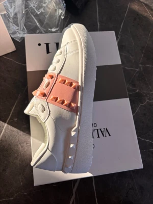 Valentino Rockstud sneakers vit/rosa - Snygga Valentino Garavani Rockstud sneakers i vitt skinn med rosa band och ikoniska nitar. Skorna har rund tå, snörning och platt sula. Perfekt statement-piece för dig som gillar exklusiva detaljer och trendig design.