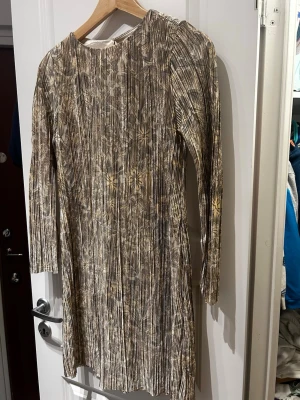 Plisserad blommig klänning från H&M - Säljer en plisserad kortklänning från H&M i beige och bruna toner med blommigt mönster. Klänningen har lång ärm, rund halsringning och glansigt material som ger en lyxig känsla. Perfekt för dig som vill sticka ut med en unik look! Svårt att se färgen då den är fotad med blixt men första bilden är mer hur den ser ut i verkligheten!