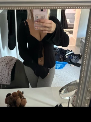 Svart långärmad knytblus - Säljer en svart långärmad blus med djup urringning och knyt framtill. Toppen är croppad och har vida ärmar som ger en snygg siluett. Perfekt för dig som gillar trendiga detaljer och vill ha något som sticker ut.