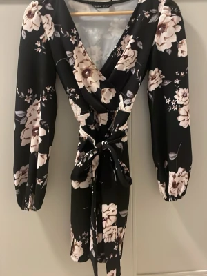 Blommig omlottklänning från SHEIN - Supersnygg kort omlottklänning från SHEIN i svart med stora beige och vita blommor. Klänningen har långa ärmar med resår vid ärmslut, v-ringning och ett knytband i midjan som ger en fin siluett. Perfekt för dig som vill ha en feminin och trendig look.