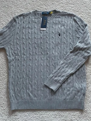 Snygg grå stickad tröja från Polo Ralph Lauren - Snygg grå kabelstickad tröja från Polo Ralph Lauren med klassisk rund halsringning och broderad logga på bröstet. Tröjan har långa ärmar och ribbade muddar vid ärmslut och nederkant. Perfekt för dig som gillar stilren och tidlös design.