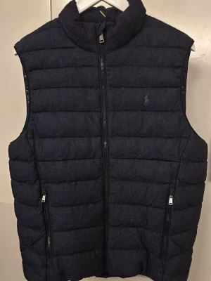 Polo Ralph Lauren dunväst (Nyskick) - Snygg mörkblå dunväst från Polo Ralph Lauren med quiltad design och hög krage. Västen har dragkedja framtill, två sidofickor med dragkedja och diskret logga på bröstet. Perfekt lager-på-lager-plagg för kyligare dagar.