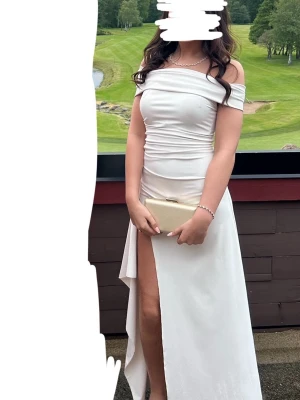 Vit offshoulder långklänning med slits - Säljer en elegant vit långklänning med offshoulder-design och hög slits framtill. Klänningen har figurnära passform och draperade detaljer över livet som ger en snygg siluett. Perfekt för bal eller student. Materialet känns mjukt och följsamt. 