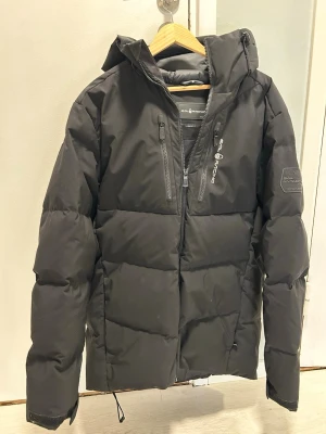 Svart dunjacka från Sail Racing, som ny! Strlk M  - Säljer en svart vinterjacka från Sail Racing med huva och dragkedjor på bröstet. Jackan har en clean look med logga på ärmen och bröstet,  Jackan är som ny, knappt använd. Storlek M 
