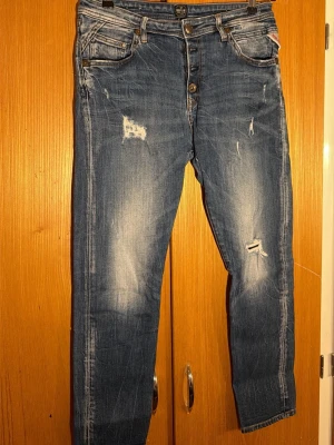  Replay jeans med slitningarblå denim - Säljer ett par blå Replay jeans med snygga slitningar och distressed detaljer framtill. Klassisk femficksmodell med knäppning i gylfen och coola kontrastsömmar. Jeansen har en rak passform och är tillverkade i tåligt denimtyg. Perfekta för dig som gillar en avslappnad streetstil.