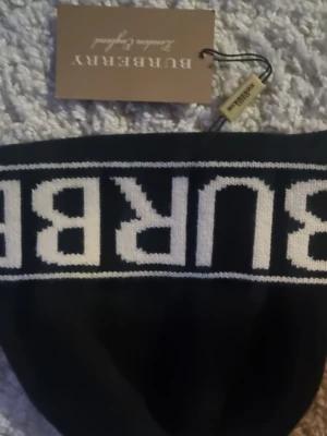 Svart mössa  från Burberry - Svart mössa från Burberry med vit logotyp i bred rand över bröstet. Klassisk och stilren design med tydlig märkning. Perfekt för dig som vill ha en snygg och enkel statement-piece i garderoben. Helt ny med peis5