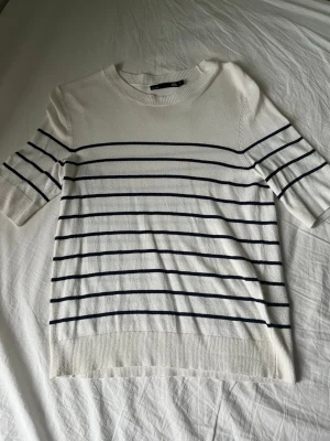 Vit randig tröja från 157 - Vit tröja från 157 med svarta horisontella ränder och rund hals. Tröjan har halvlånga ärmar och är gjord i ett mjukt, stickat material. Perfekt för dig som gillar en clean och klassisk look.