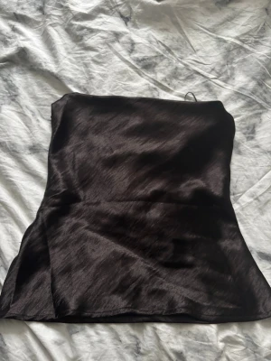Svart satin tubtopp från Gina Tricot - Säljer en svart tubtopp i glansigt satinmaterial från Gina Tricot. Toppen har smala snören som kan knytas i ryggen för en snygg och justerbar passform. Perfekt för dig som gillar trendiga och stilrena plagg med en elegant touch.Den är från Gina i storlek 40
