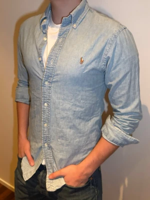 Ljusblå skjorta från Ralph Lauren - Säljer en klassisk ljusblå skjorta från Ralph Lauren med button-down krage och broderad logga på bröstet. Skjortan har lång ärm, normal passform och är tillverkad i mjuk bomull. Perfekt att bära öppen över en t-shirt för en avslappnad look.